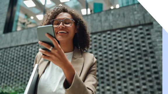 Person in beige blazer holding a laptop and looking at their smartphone outside a modern building.