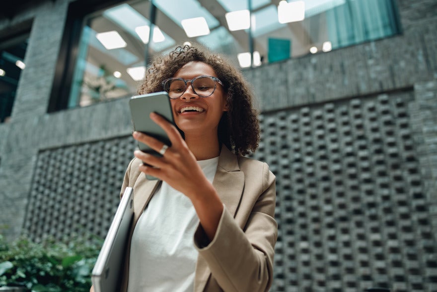 Person in beige blazer holding a laptop and looking at their smartphone outside a modern building.