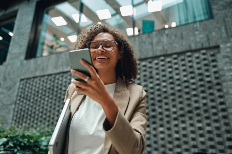 Person in beige blazer holding a laptop and looking at their smartphone outside a modern building.
